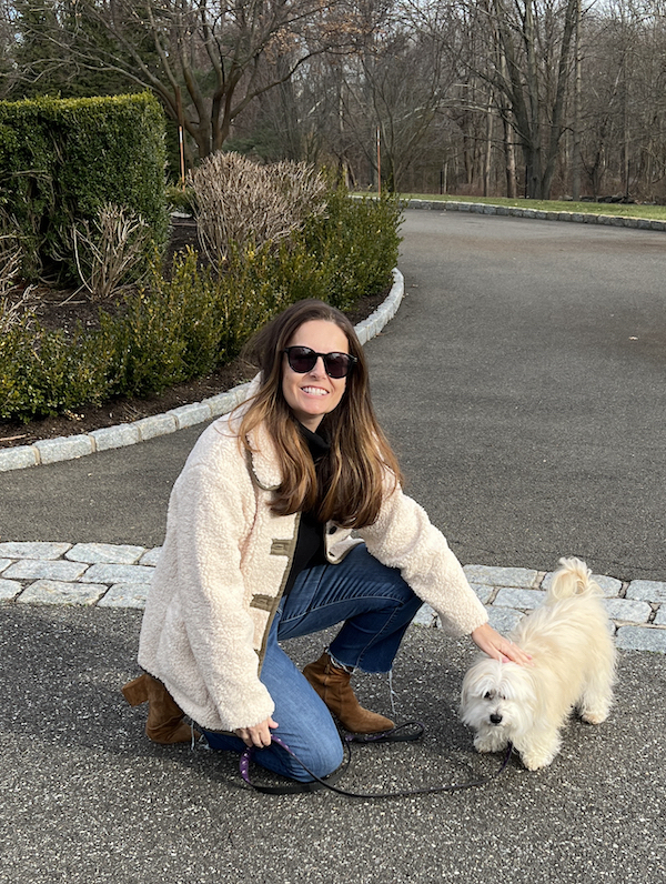 woman with small white dog on leash bending down wearing a white fluffy fleece coat.