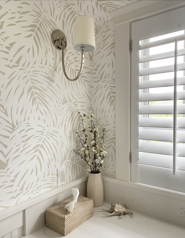 corner of beach cottage bathroom with white wood blinds and brass sconce.